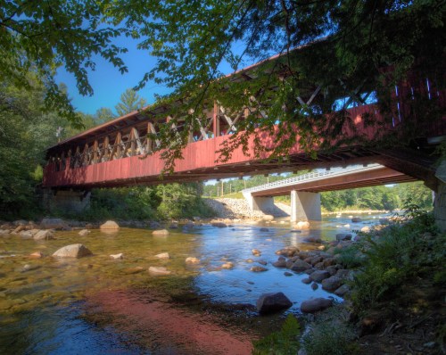 A red covered bridge spans a river, surrounded by trees and rocky banks under a clear blue sky.
