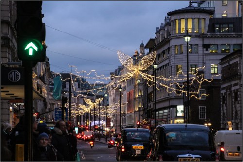A city street adorned with festive lights, including angel designs, under a twilight sky with traffic and pedestrians.