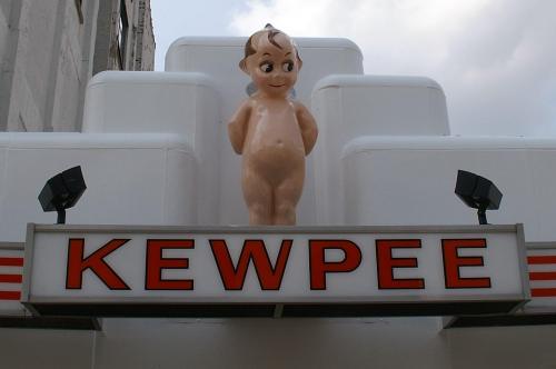 A large, smiling baby figure stands atop a sign that reads "KEWPEE," against a cloudy sky.