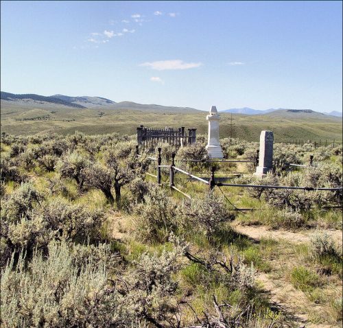 A remote graveyard surrounded by sagebrush, featuring two white tombstones and a wooden fence against a mountainous backdrop.