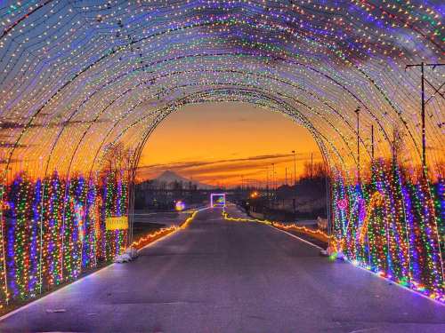 A colorful light tunnel arches over a road, with a sunset and mountains in the background.
