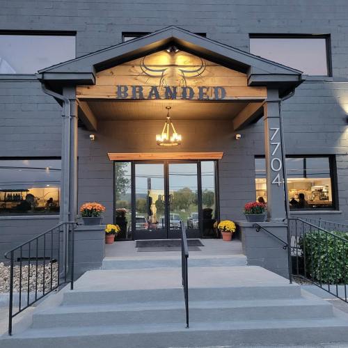 Entrance of a building with the sign "BRANDED," featuring a porch, steps, and potted flowers on either side.