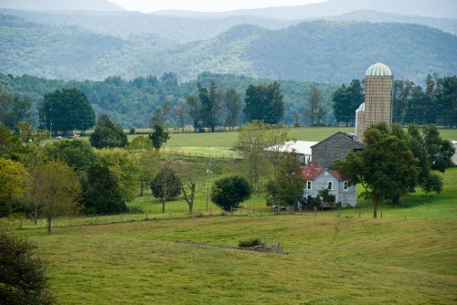 A scenic view of a farm with a barn and silo, surrounded by lush green fields and distant mountains.