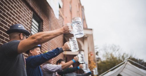 A group of people raising large beer mugs outdoors, celebrating with smiles on a cloudy day.