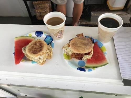 Two breakfast sandwiches on paper plates with watermelon slices, accompanied by cups of coffee.
