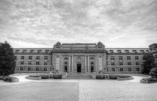 Historic building with ornate architecture, surrounded by trees and a cloudy sky, in black and white.