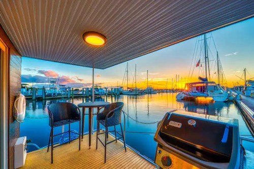 A scenic marina view at sunset, featuring boats, a grill, and a small table with chairs on a deck.