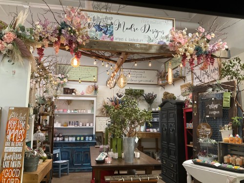 A cozy shop interior filled with floral decor, candles, and handmade items, featuring warm lighting and rustic furniture.