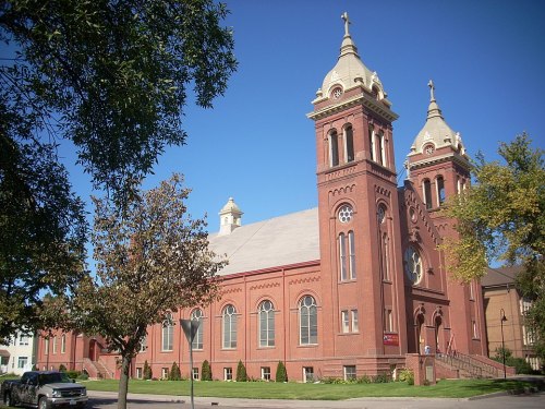 A red brick church with two tall towers and a clear blue sky in the background, surrounded by trees.