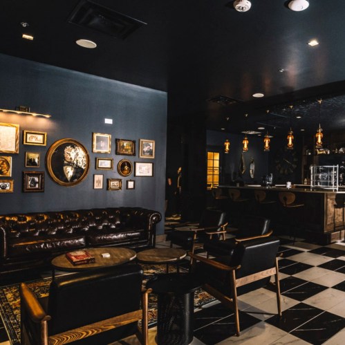 A stylish lounge with dark walls, vintage decor, a leather sofa, and a bar area featuring pendant lights and checkered flooring.
