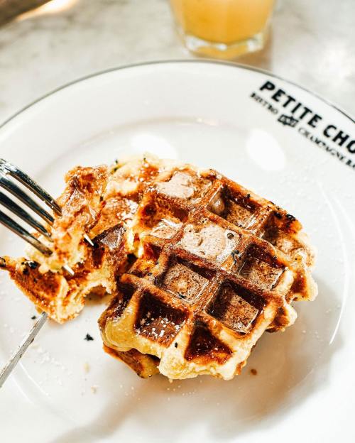 A fork holding a piece of golden-brown waffle, dusted with powdered sugar, on a white plate with "Petite Chou" printed on it.