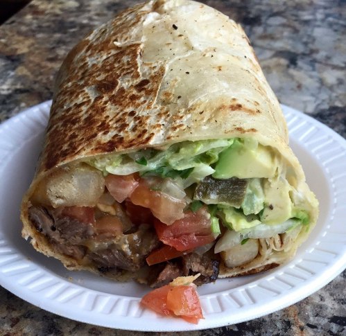 A close-up of a grilled burrito filled with meat, lettuce, tomatoes, and avocado, served on a white plate.