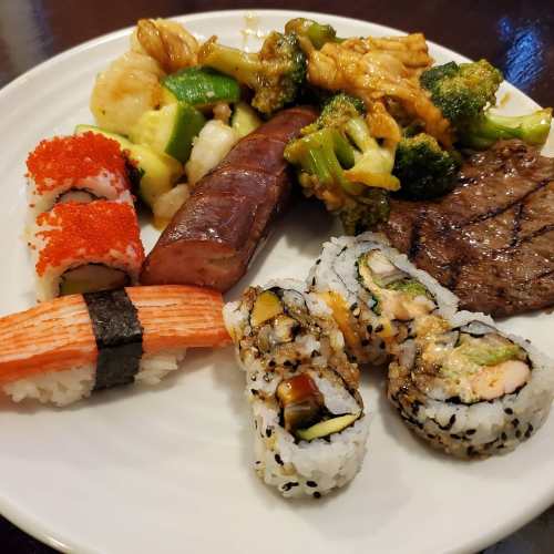 A plate of sushi, grilled steak, sausage, and mixed vegetables, featuring colorful rolls and seafood.