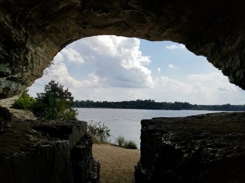 View through a rocky cave opening, revealing a river and cloudy sky in the background.