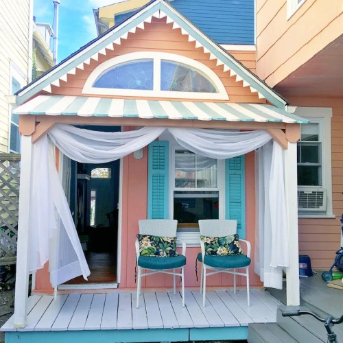 A charming porch with two floral chairs, sheer white curtains, and colorful siding on a cozy house.