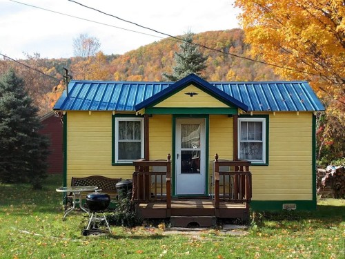 A small yellow house with a blue metal roof, surrounded by autumn trees and a grassy yard.