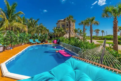 A sunny poolside scene with a blue pool, lounge chairs, a hammock, and palm trees, set against a clear blue sky.
