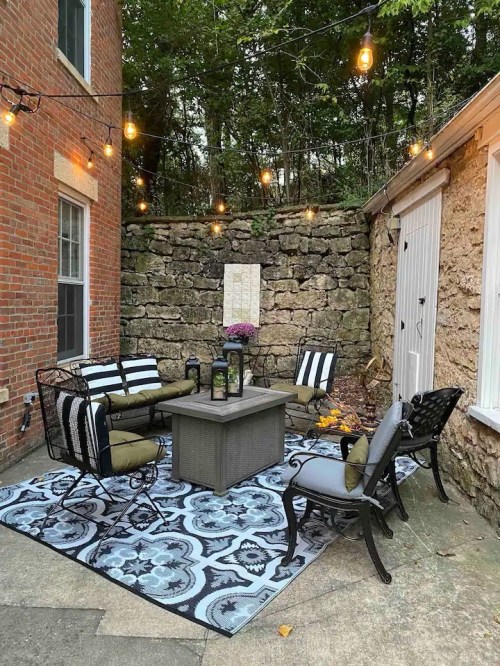 Cozy outdoor seating area with string lights, a patterned rug, and a fire pit surrounded by stone walls and greenery.