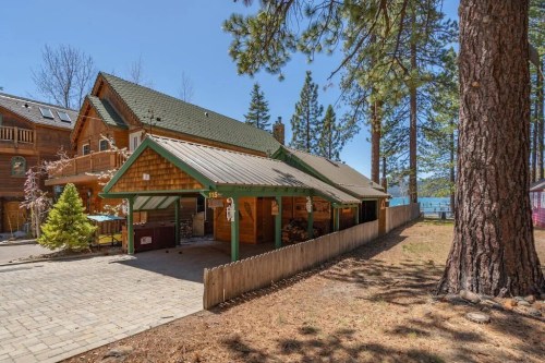 A cozy wooden cabin with a green roof, surrounded by tall trees and a clear blue sky, near a lake.