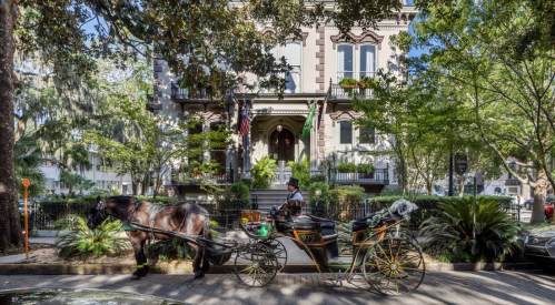 A horse-drawn carriage is parked in front of a historic building surrounded by lush greenery and trees.