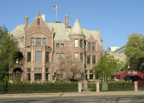 A large, historic mansion with a turret and a flag, surrounded by greenery and a decorative fence.