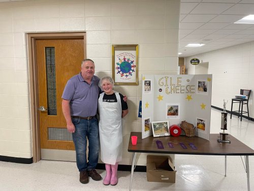 A man and a woman stand beside a display board about cheese in a school hallway.
