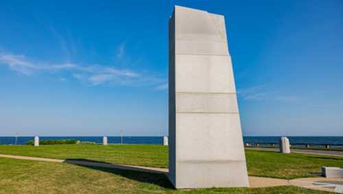 A tall, angled stone monument stands on a grassy area near the ocean under a clear blue sky.