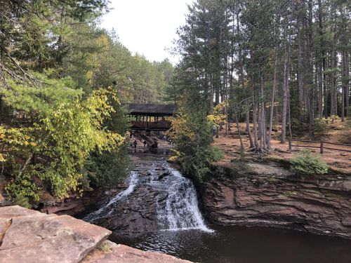 A serene waterfall flows over rocks, surrounded by trees and a rustic covered bridge in a peaceful forest setting.