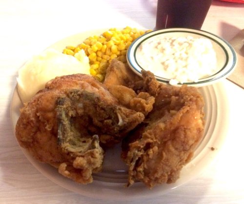 A plate of fried chicken with corn, mashed potatoes, and a side of cottage cheese. A drink is visible in the background.