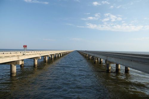 A long bridge stretches over calm water, disappearing into the horizon under a clear blue sky.