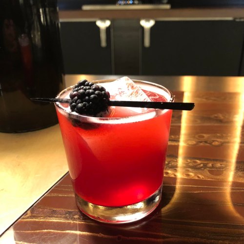 A vibrant red cocktail with ice, garnished with a blackberry and served in a short glass on a wooden table.