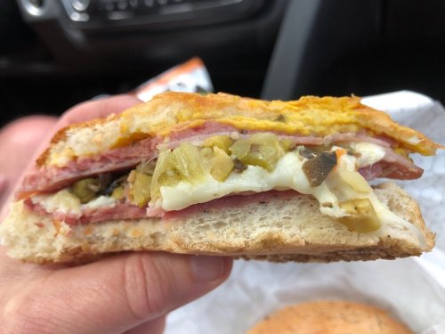 A close-up of a hand holding a sandwich filled with layers of meat, cheese, and pickles, with a toasted bun.