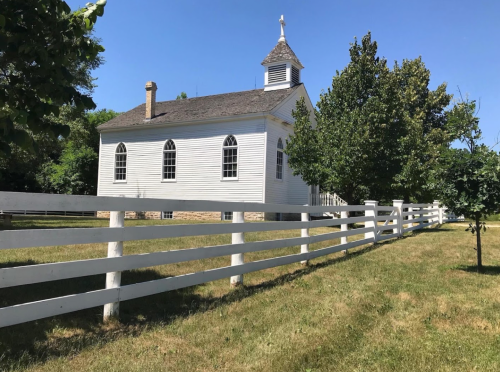 A white, historic building with arched windows, surrounded by green grass and trees, next to a white fence under a clear blue sky.