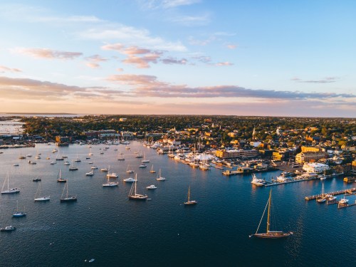 Aerial view of a harbor at sunset, filled with sailboats and surrounded by a picturesque coastal town.