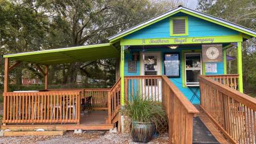 A colorful bagel shop with a wooden deck and awning, surrounded by trees and outdoor seating.