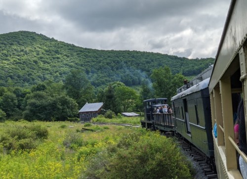 A vintage train travels through lush green hills, with passengers enjoying the scenic view from the open car.