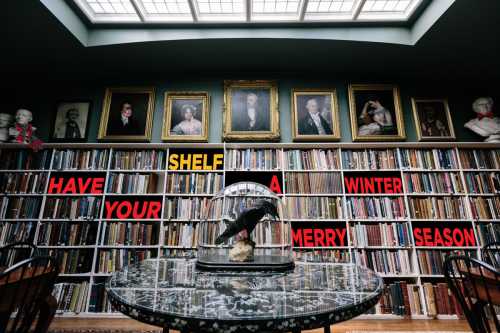 A cozy library with a large bookshelf, a glass display case, and festive text on the wall wishing a merry winter season.