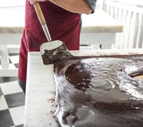 A person in an apron spreads melted chocolate on a marble surface with a spatula.