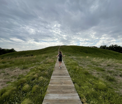 A person walks along a wooden path leading to a grassy hill under a cloudy sky.