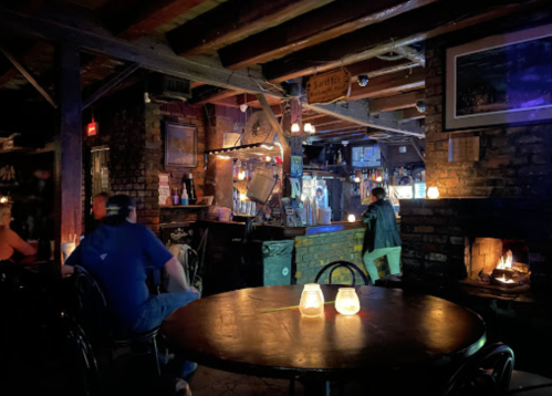 A dimly lit bar with wooden beams, candles on tables, and patrons enjoying the cozy atmosphere.