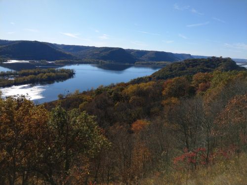 A scenic view of a river surrounded by hills and colorful autumn foliage under a clear blue sky.