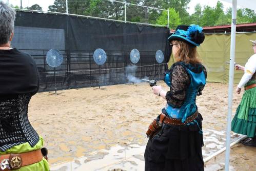 A woman in a blue corset and hat fires a gun at targets in a shooting range, surrounded by others in period costumes.