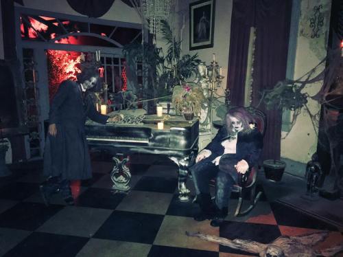A dimly lit room with eerie decorations, featuring a piano and two ghostly figures, one seated and one leaning over the piano.
