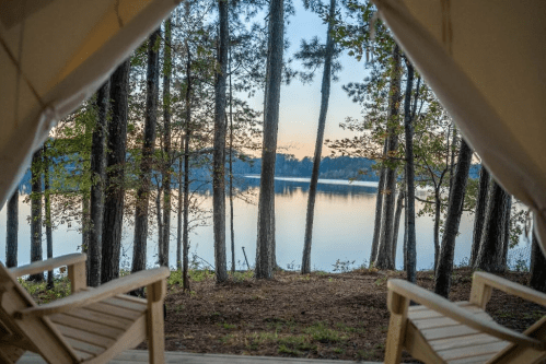 View from a tent overlooking a calm lake surrounded by tall trees at sunrise. Two wooden chairs in the foreground.