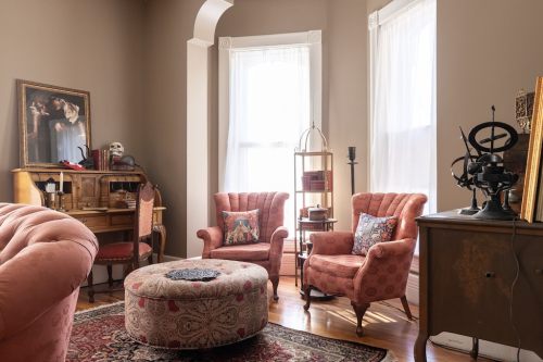 Cozy room with two pink armchairs, a round ottoman, a wooden desk, and vintage decor against warm-toned walls.