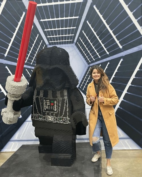 A woman stands next to a large LEGO Darth Vader figure, holding a red lightsaber, in a themed background.