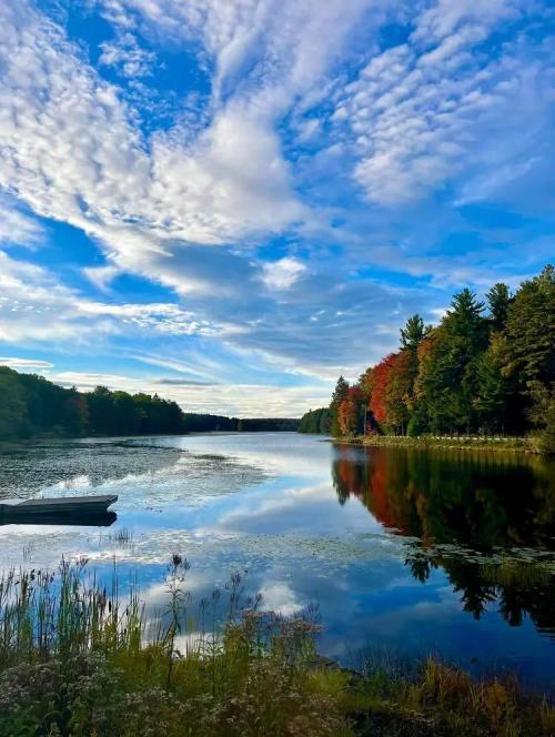 A serene lake reflects a blue sky with clouds, surrounded by trees showing autumn colors. A small boat is visible on the shore.