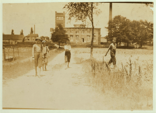 Four boys in overalls walking along a dirt path, with a factory and trees in the background, in a vintage sepia tone.