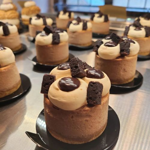 Close-up of mini chocolate mousse cakes topped with cream, chocolate pieces, and cookie crumbles on black bases.