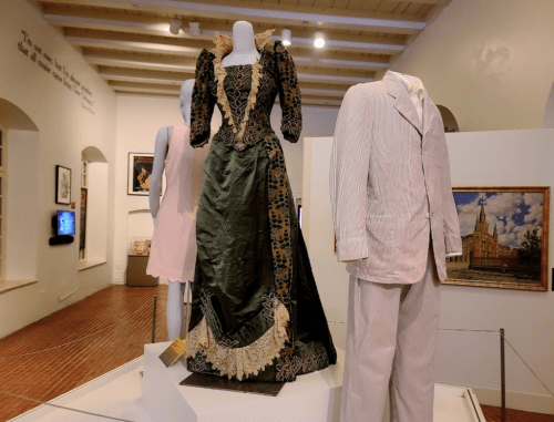 A vintage green gown with lace details and a light-colored suit displayed in a museum setting.